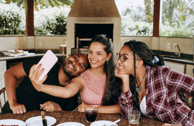 Three friends taking a selfie together at a table with food outdoors, capturing a joyful moment.