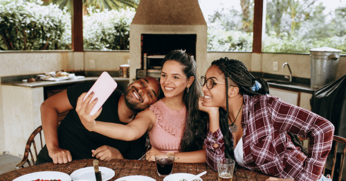 Three friends taking a selfie together at a table with food outdoors, capturing a joyful moment.