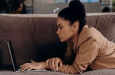 African American woman lying on sofa working remotely on her laptop