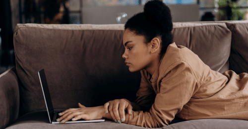 African American woman lying on sofa working remotely on her laptop