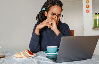 A woman concentrates while working on her laptop at home, enjoying coffee and bread